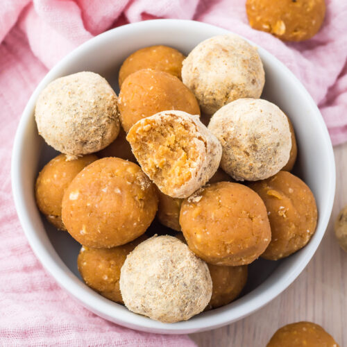 A white bowl filled with peanut butter protein balls, surrounded by a pink cloth with more peanut butter protein balls scattered on the table.