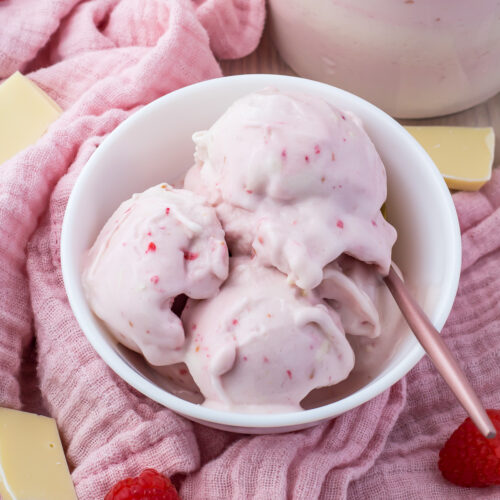 A bowl of White chocolate raspberry protein ice cream in a bowl next to a ninja creami pint and a pink cloth and an ice cream scoop. White chocolate and raspberries are scattered around.