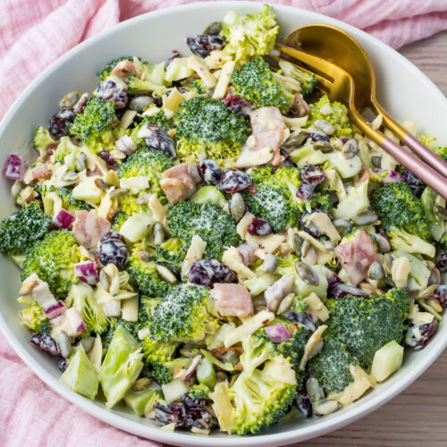 Broccoli salad in a white bowl with a pink and gold fork and spoon. A pink cloth surrounds it.