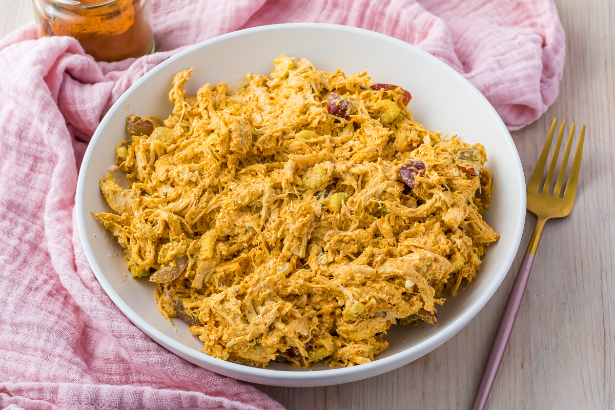 Overhead photo of a bowl of high protein curry chicken salad, on a table with a pink cloth, curry powder, and fork.