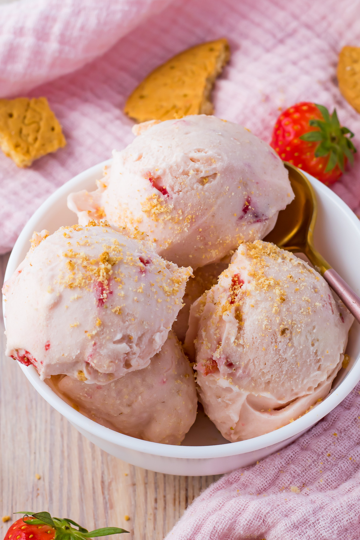 Strawberry cottage cheese ice cream in a bowl beside a pink cloth with strawberries and cookies scattered around.