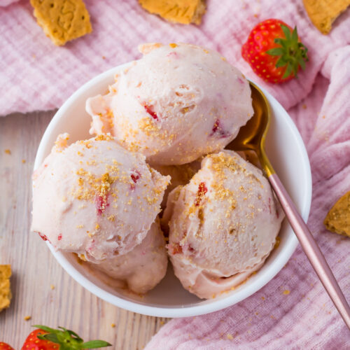 Strawberry cottage cheese ice cream in a bowl beside a pink cloth with strawberries and cookies scattered around.