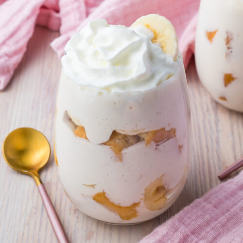 A glass filled with creamy cottage cheese banana pudding topped with whipped cream, surrounded by a gold spoon and pink fabric.