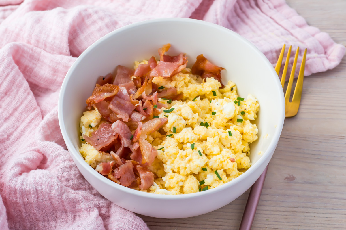 A scrambled egg cottage cheese bowl, topped with bacon bits and chives, rests on a wooden table. Nearby, a pink cloth and a gold fork add an elegant touch.