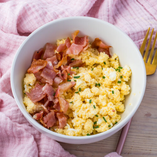 A bowl of creamy scrambled eggs mixed with cottage cheese, garnished with chives and crispy bacon pieces, sits on a wooden table beside a gold fork and a pink cloth.