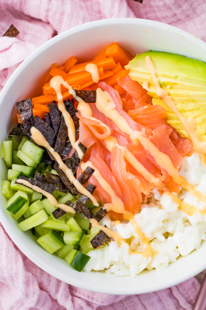 A smoked sushi bowl with smoked salmon, creamy avocado, crisp cucumber, vibrant carrots, and rice wrapped in seaweed, all topped with a flavorful drizzle of sauce.