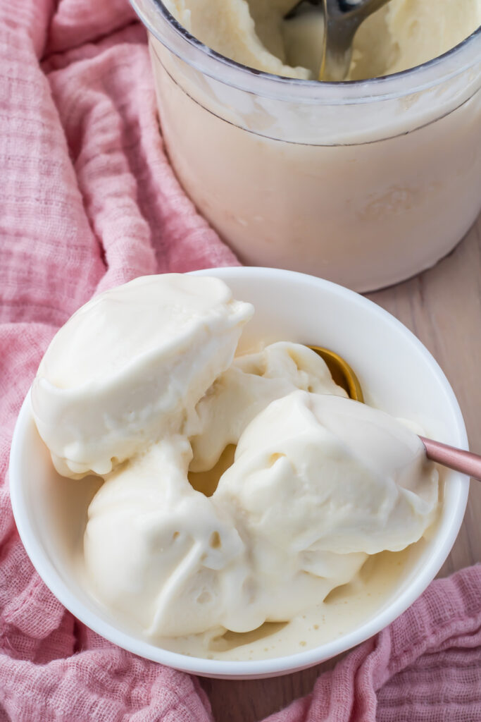 A bowl of soft-serve vanilla ice cream with a spoon rests on a wooden surface, complemented by a pink cloth.