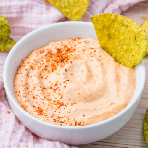 A white bowl of creamy taco dip topped with red seasoning sits alongside greenish-yellow tortilla chips, all elegantly placed on a pink cloth.