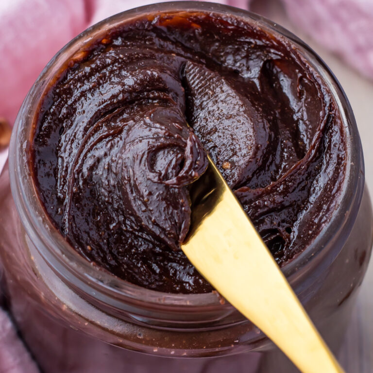 A jar filled with dark brown protein Nutella spread. A gold knife is dipped into the spread, showcasing its smooth and creamy texture, enriched with a hint of protein.