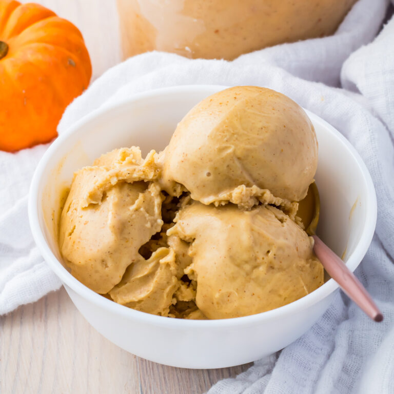 A white bowl of pumpkin ice cream with three creamy scoops sits invitingly, a spoon poised at the ready, on a light wooden surface. Nearby, a small pumpkin adds charm, hinting at the protein-packed deliciousness within each bite.