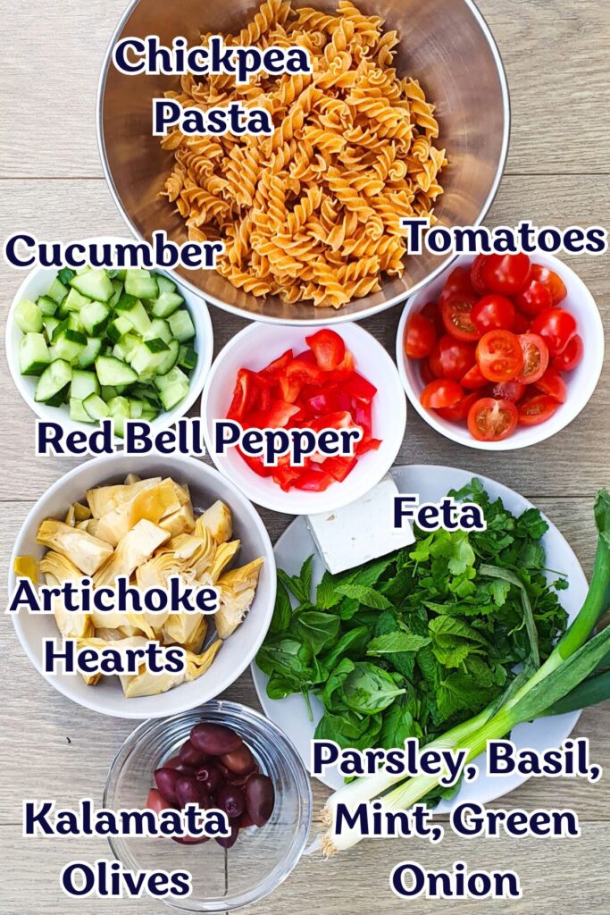 Ingredients for a high-protein pasta salad: bowls of chickpea pasta, tomatoes, cucumber, red bell pepper, artichoke hearts, feta, kalamata olives, green onion, parsley, basil, and mint on a wooden surface.