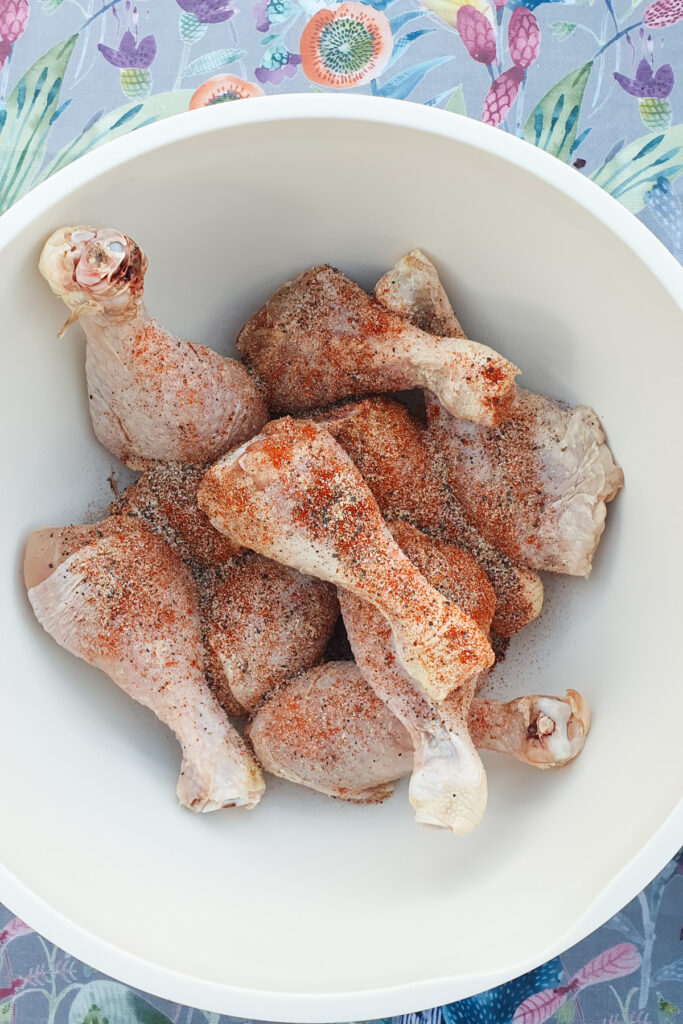 Raw chicken legs seasoned with spices rest in a white bowl.
