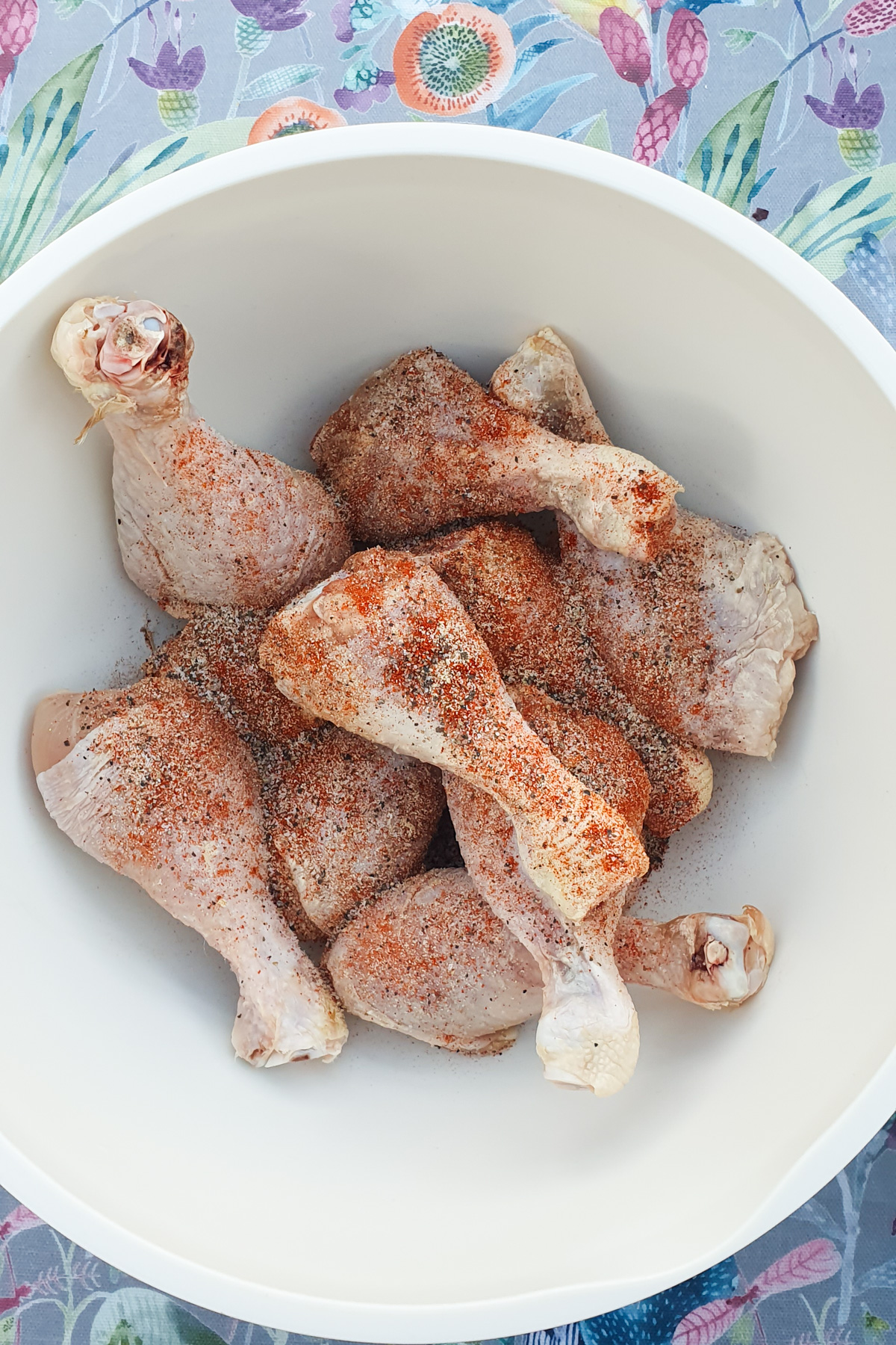 Raw chicken legs seasoned with spices rest in a white bowl.