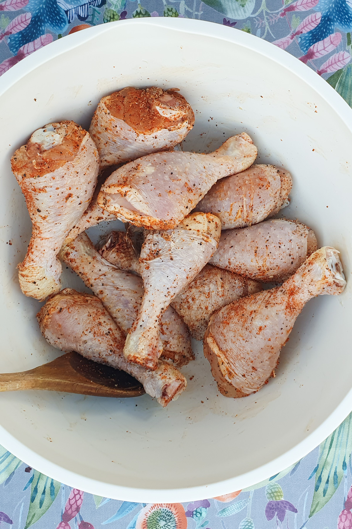 A white bowl contains several raw chicken legs seasoned with spices, accompanied by a wooden spoon.