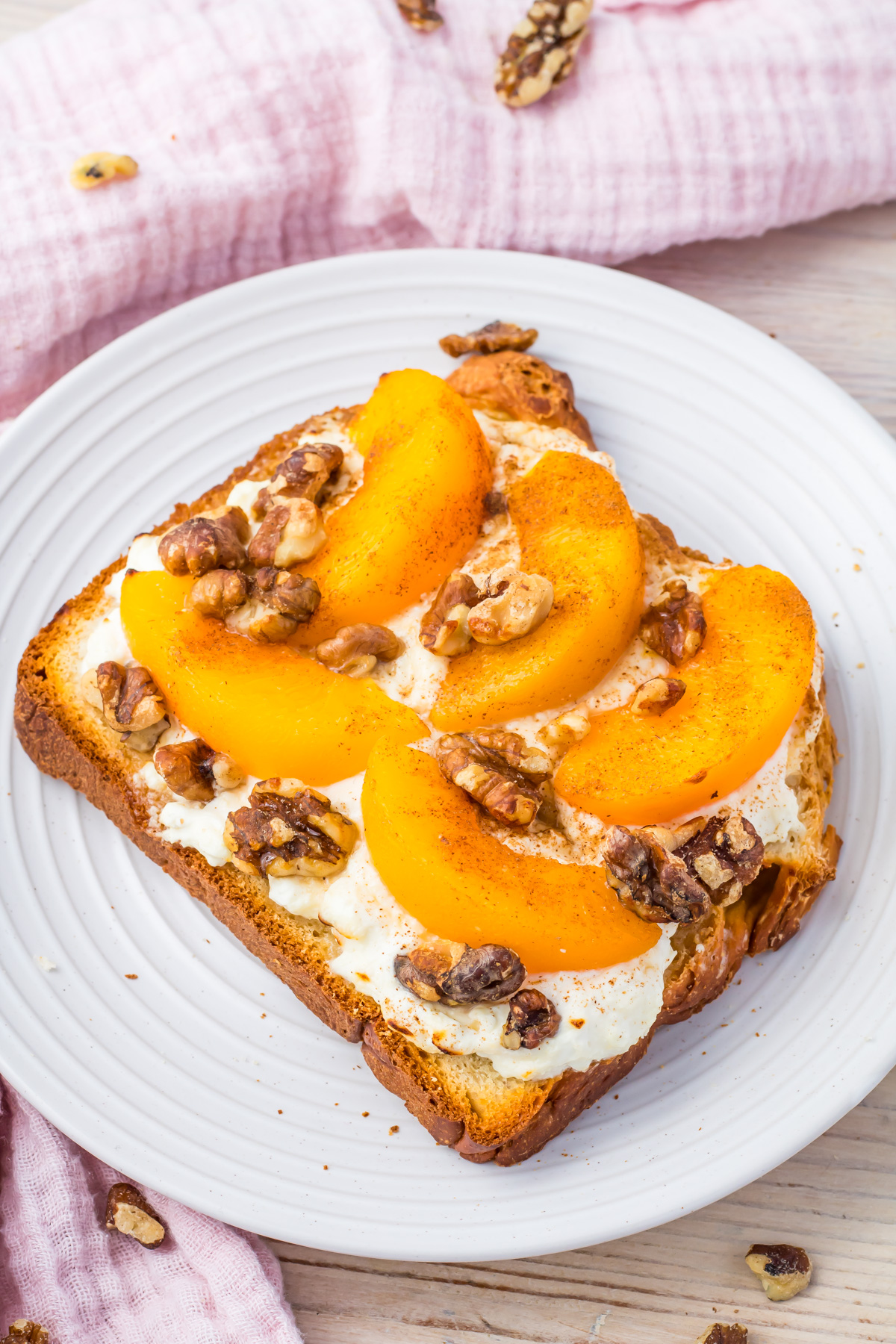 Air fryer cottage cheese toast with peach slices, and crunchy walnuts rests on a white plate atop a wooden table. A pink cloth adds a soft touch to the background.