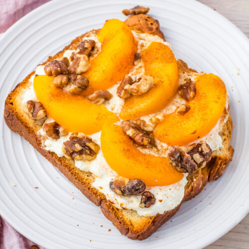 Air fryer cottage cheese toast with peach slices, and crunchy walnuts rests on a white plate atop a wooden table. A pink cloth adds a soft touch to the background.