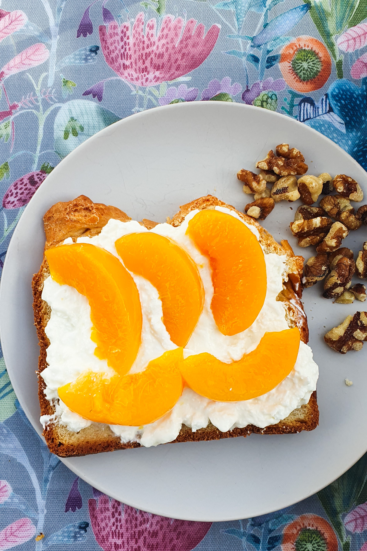 Bread topped with creamy cottage cheese and sliced peaches rests on a plate, accompanied by crunchy walnuts, all set on a floral tablecloth.