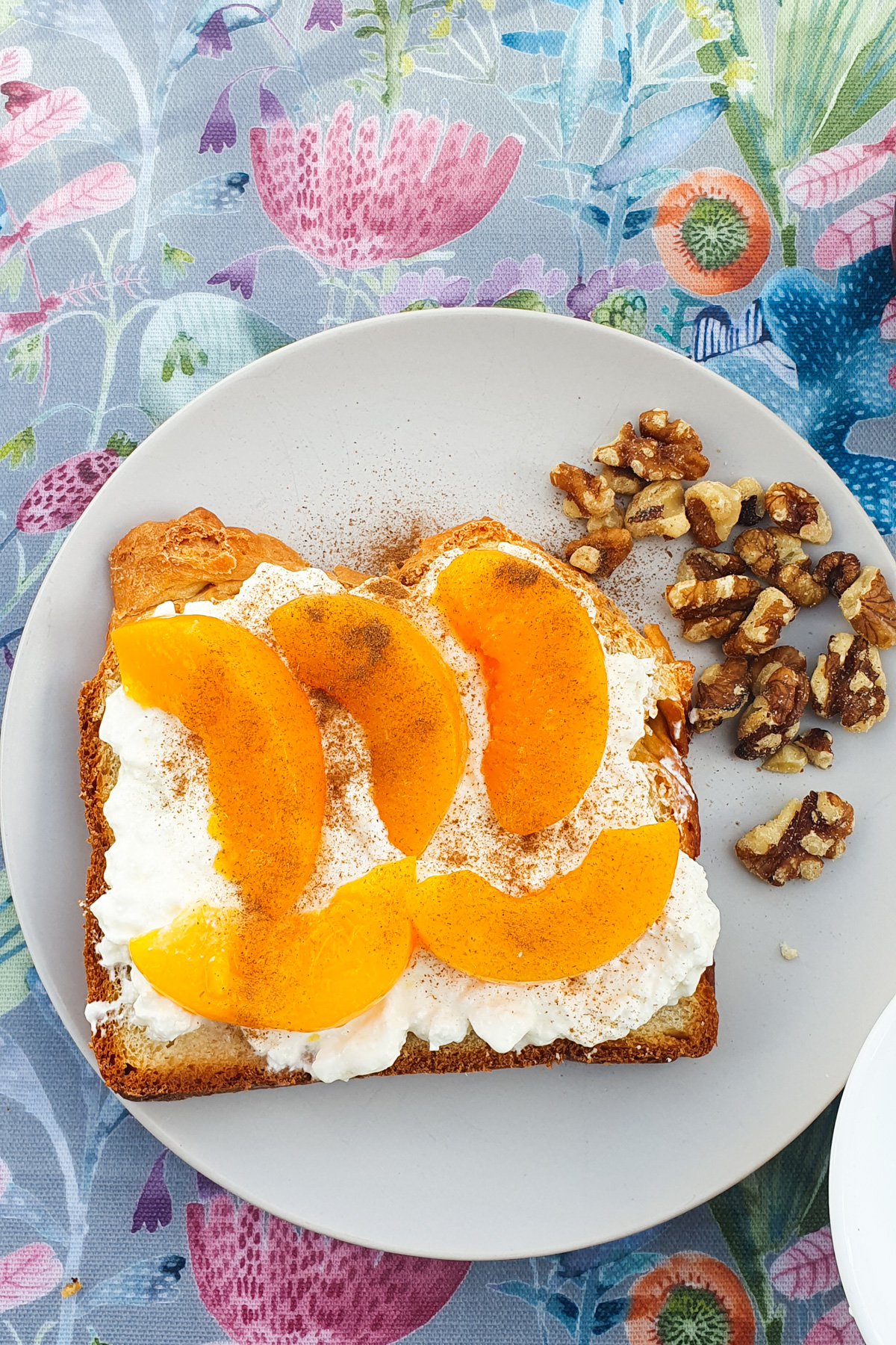 Bread topped with creamy cottage cheese and sliced peaches and cinnamon rests on a plate, accompanied by crunchy walnuts, all set on a floral tablecloth.