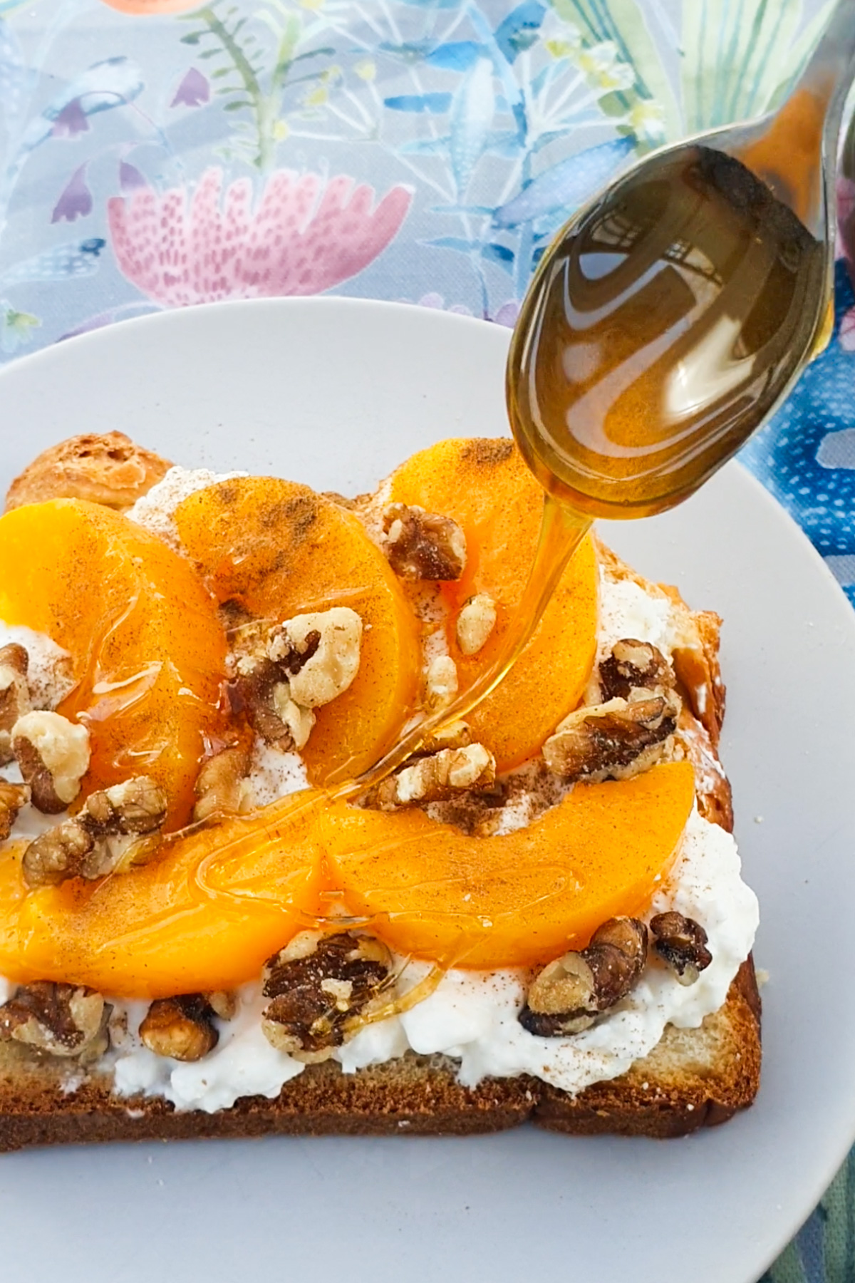 Peach slices, walnuts, and a drizzle of honey atop cottage cheese on bread, on a white plate, set against a floral tablecloth.