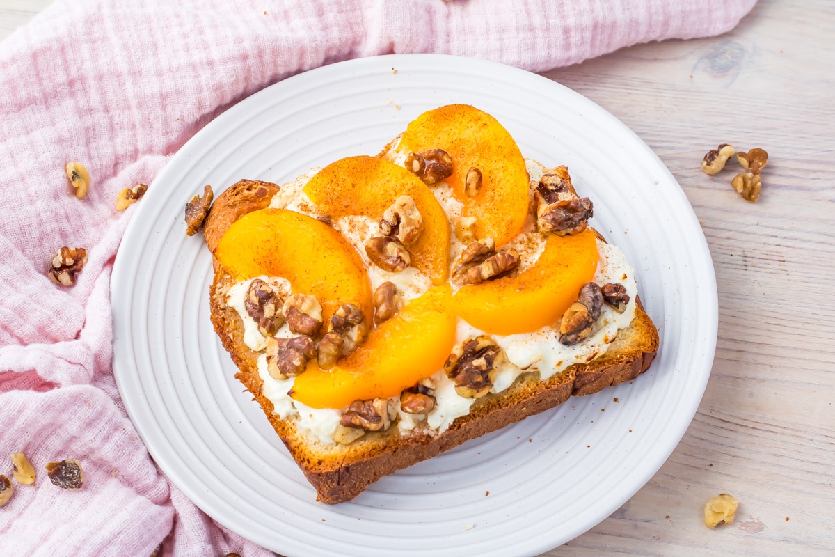 Air fryer cottage cheese toast with peach slices, and crunchy walnuts rests on a white plate atop a wooden table. A pink cloth adds a soft touch to the background.
