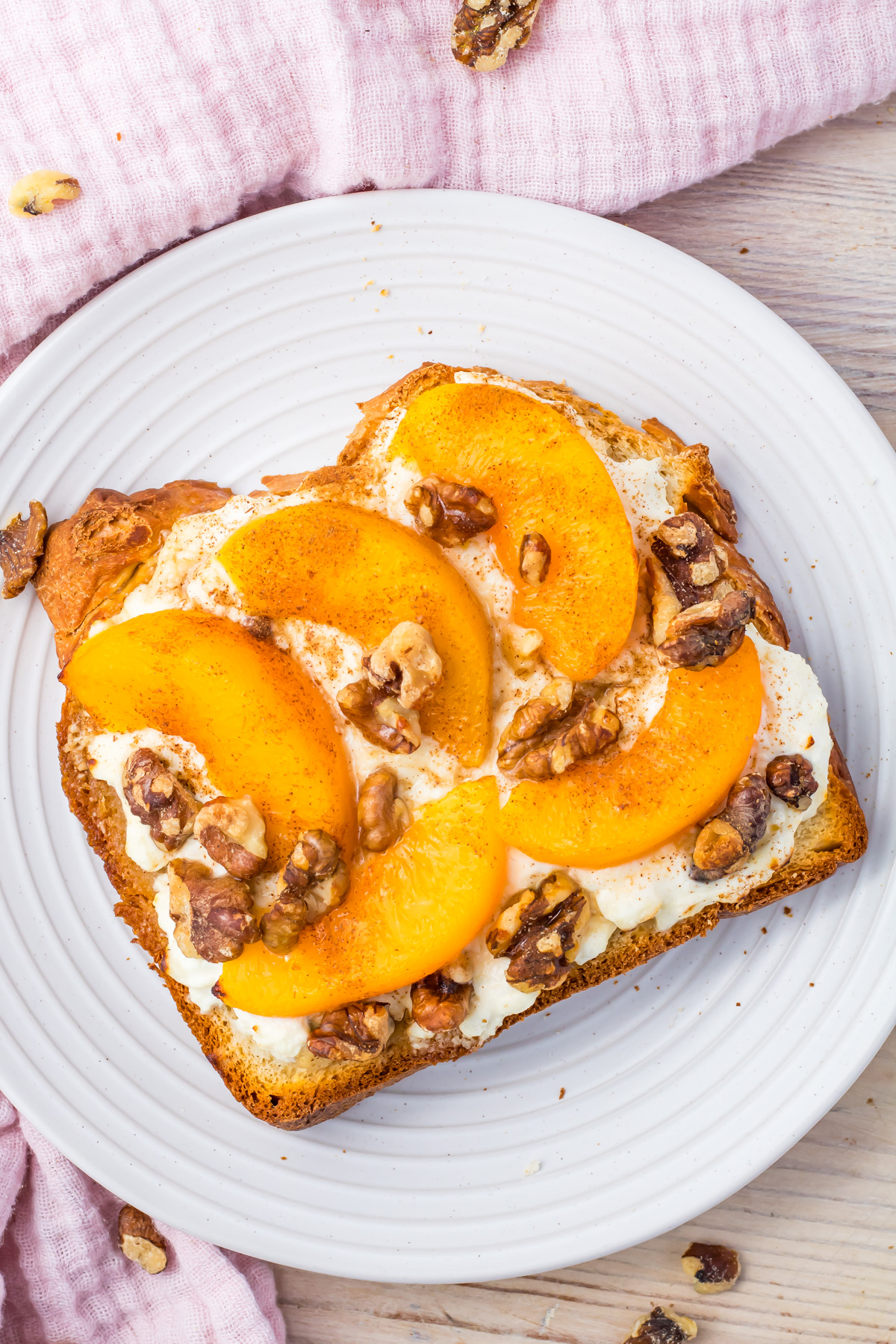 Air fryer cottage cheese toast with peach slices, and crunchy walnuts rests on a white plate atop a wooden table. A pink cloth adds a soft touch to the background.
