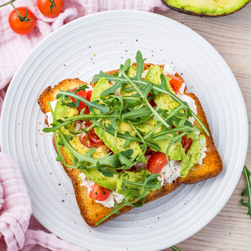 A white plate holds toast adorned with avocado slices, creamy cottage cheese, cherry tomatoes, and a sprinkle of arugula.