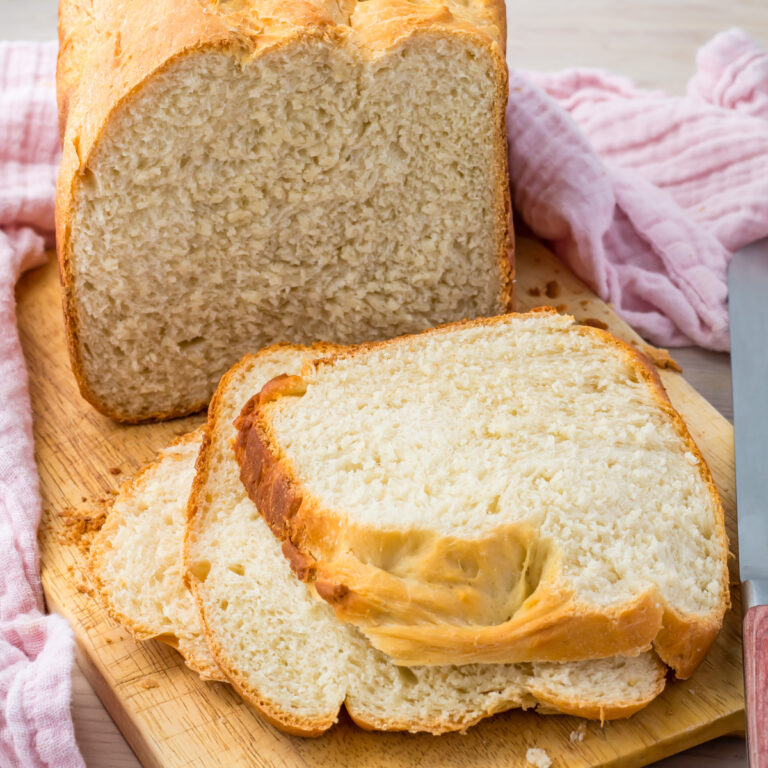 A freshly baked loaf of bread machine cottage cheese bread, along with its slices, rests invitingly on a wooden cutting board. A trusty knife lies next to it, accompanied by a soft pink cloth.
