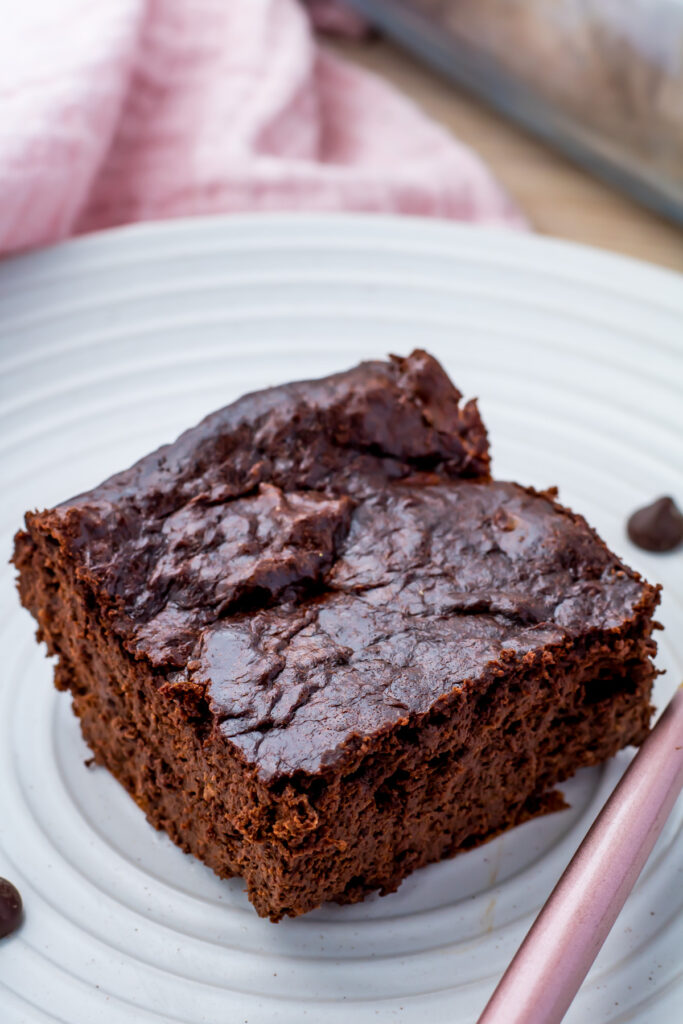 A rich square of cottage cheese brownie rests on a white plate, beside a whimsical pink utensil and cloth.