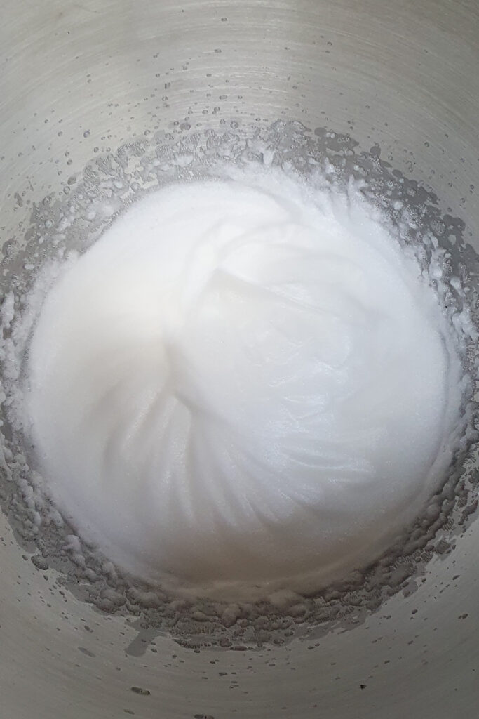 A bowl of whipped egg whites forming soft peaks in a metallic mixing bowl.