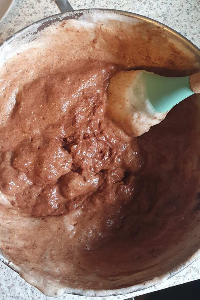 A green spatula blends chocolate brownie batter with egg whites in a metal bowl on a speckled countertop.