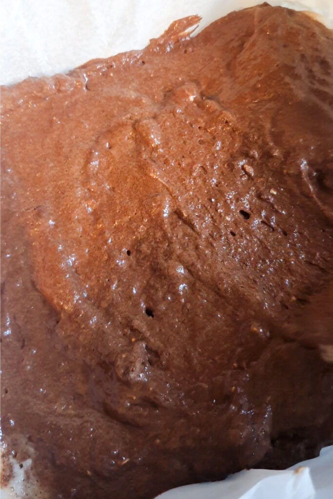 A close-up of uncooked, glossy brownie batter in a baking tray lined with parchment paper.