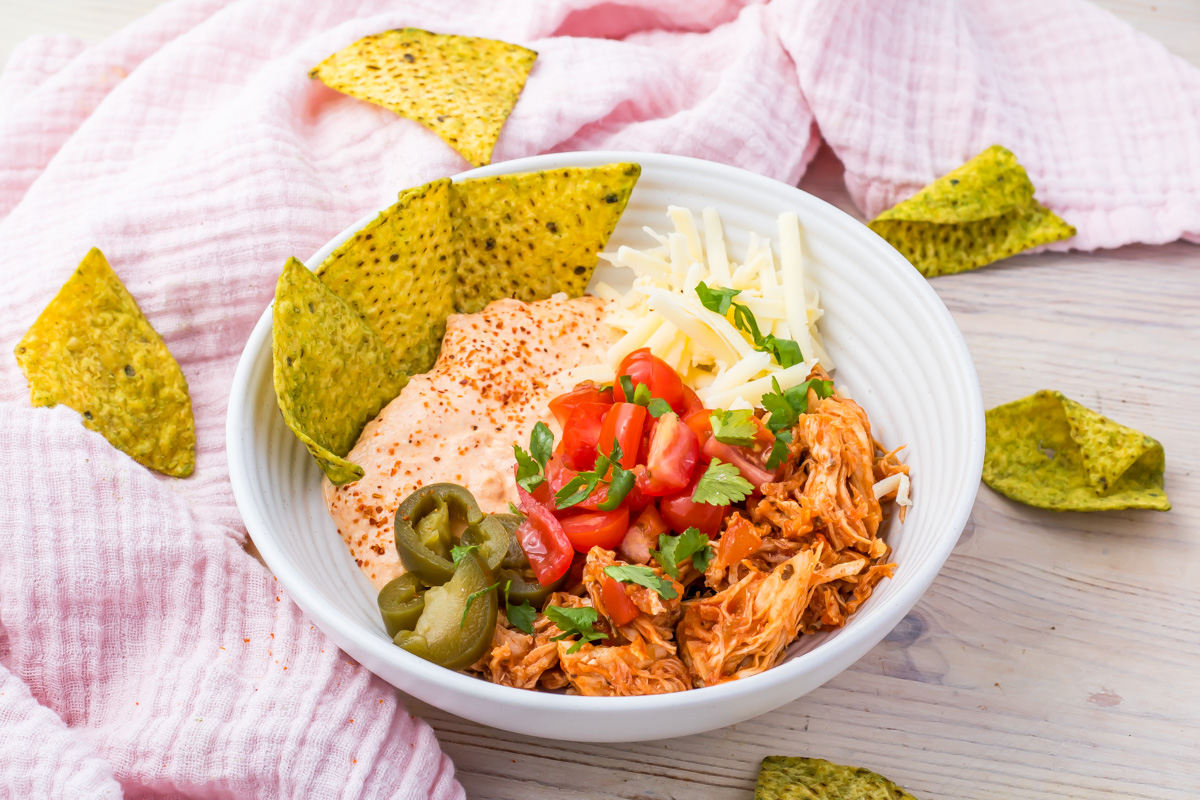 A white Cottage Cheese Chicken Taco Bowl brimming with shredded chicken, sliced jalapeños, diced tomatoes, and cheese pairs perfectly with chips on a wooden surface.