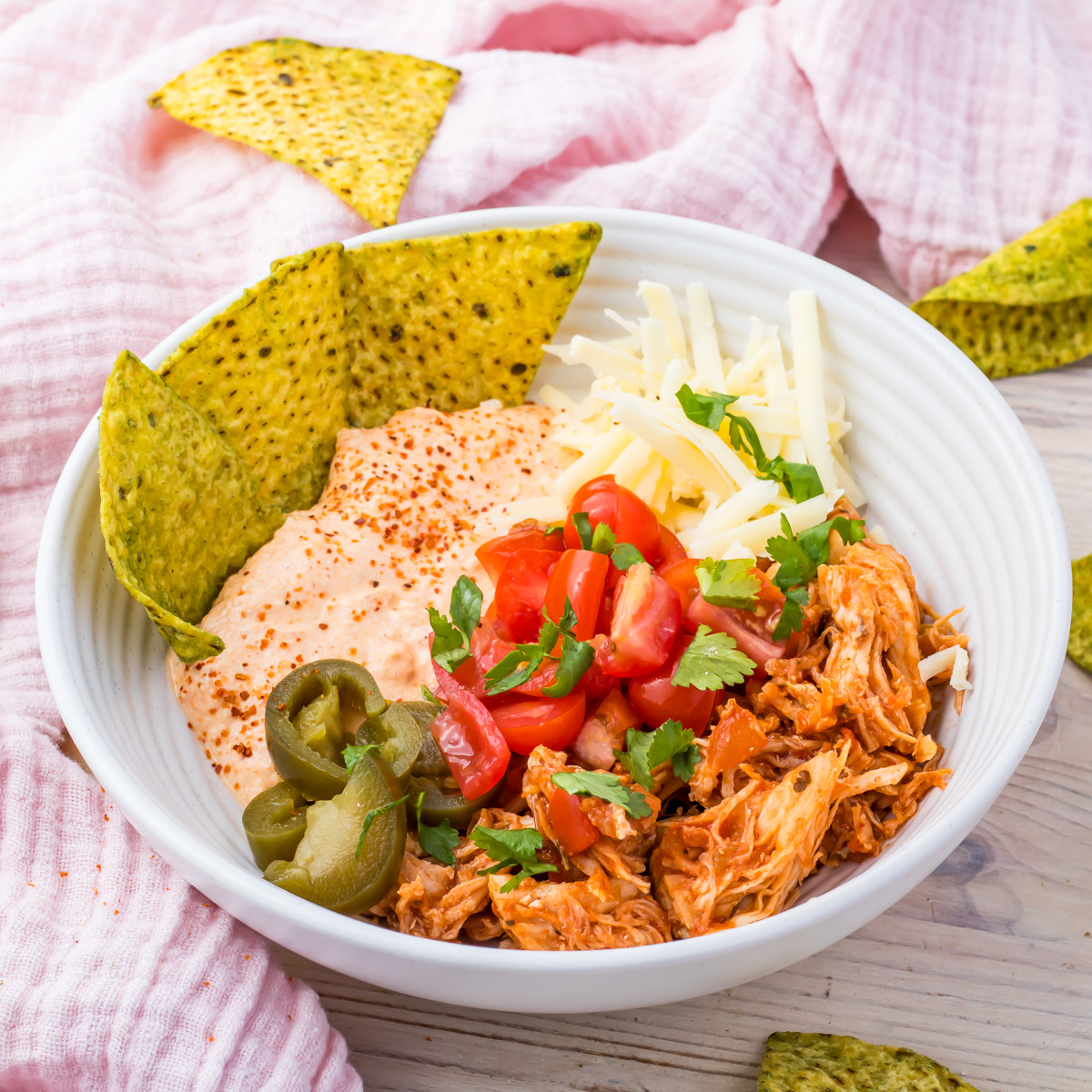 A white Cottage Cheese Chicken Taco Bowl brimming with shredded chicken, sliced jalapeños, diced tomatoes, and cheese pairs perfectly with chips on a wooden surface.