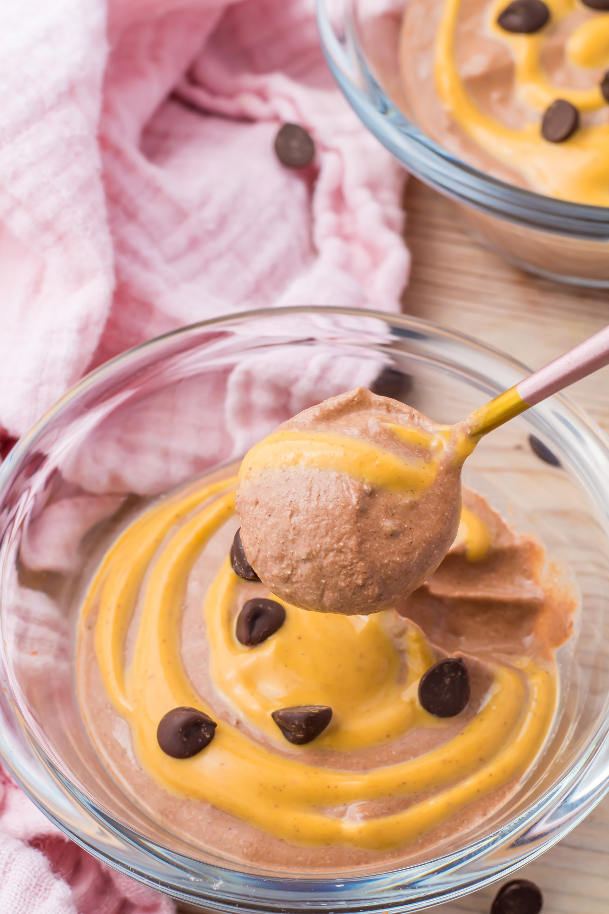 A bowl of chocolate cottage cheese mousse topped with peanut butter drizzle and chocolate chips, with a spoon holding a creamy scoop. A hint of pink cloth is partially visible in the background.
