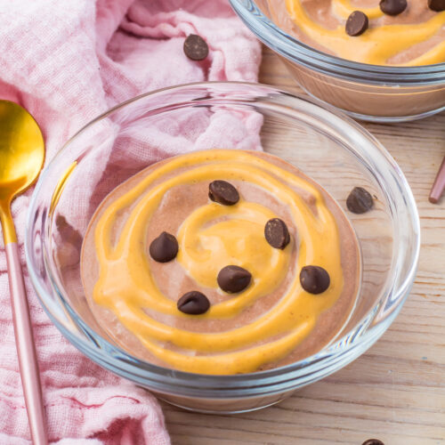 Glass bowl featuring cottage cheese mousse elegantly swirled with peanut butter and topped with chocolate chips. A pink cloth and gold spoon provide a charming background.