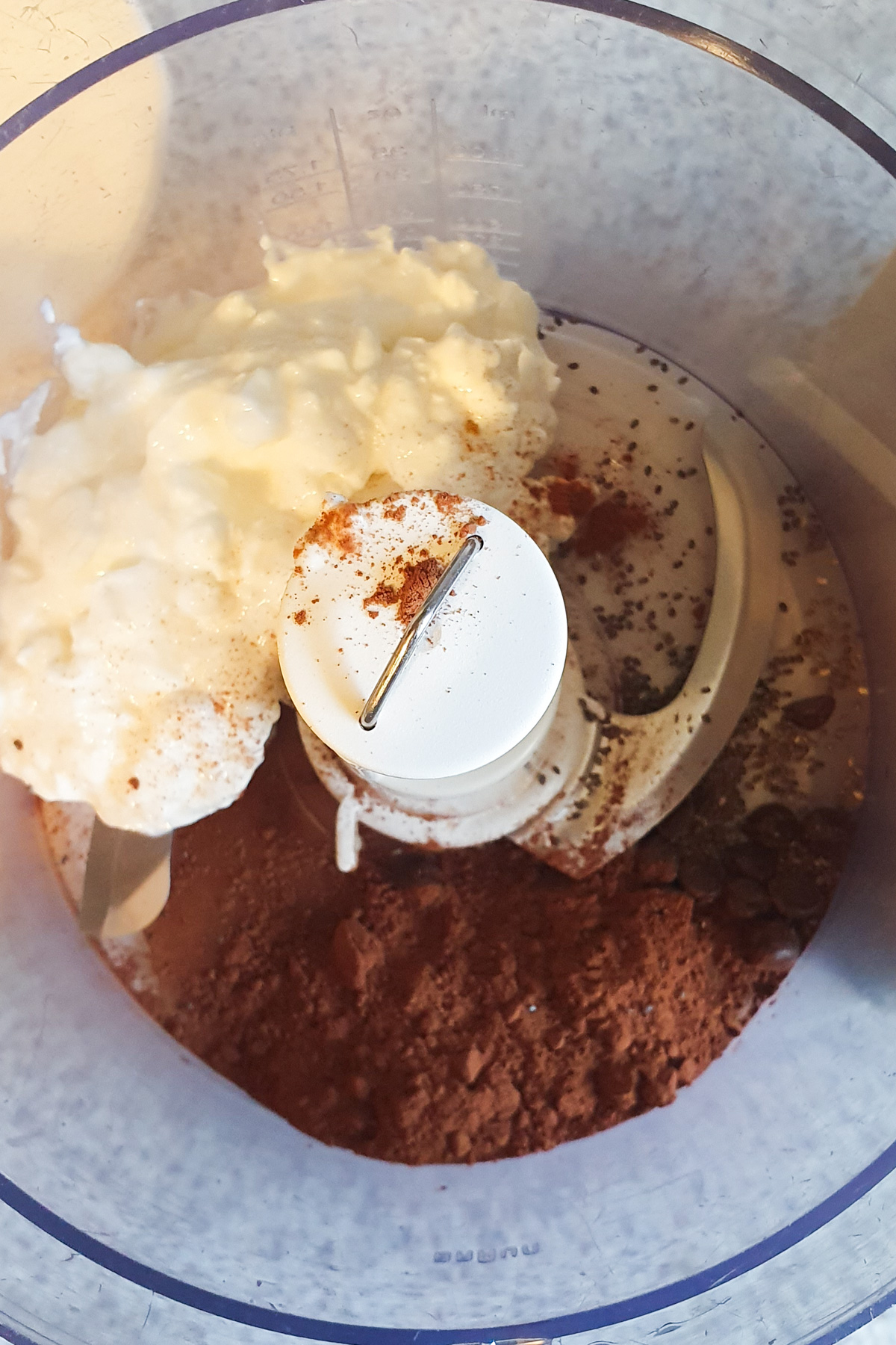 A food processor bowl filled with cottage cheese, cocoa powder, chia seeds, and a sprinkling of instant coffee is ready for blending into a luscious mousse.