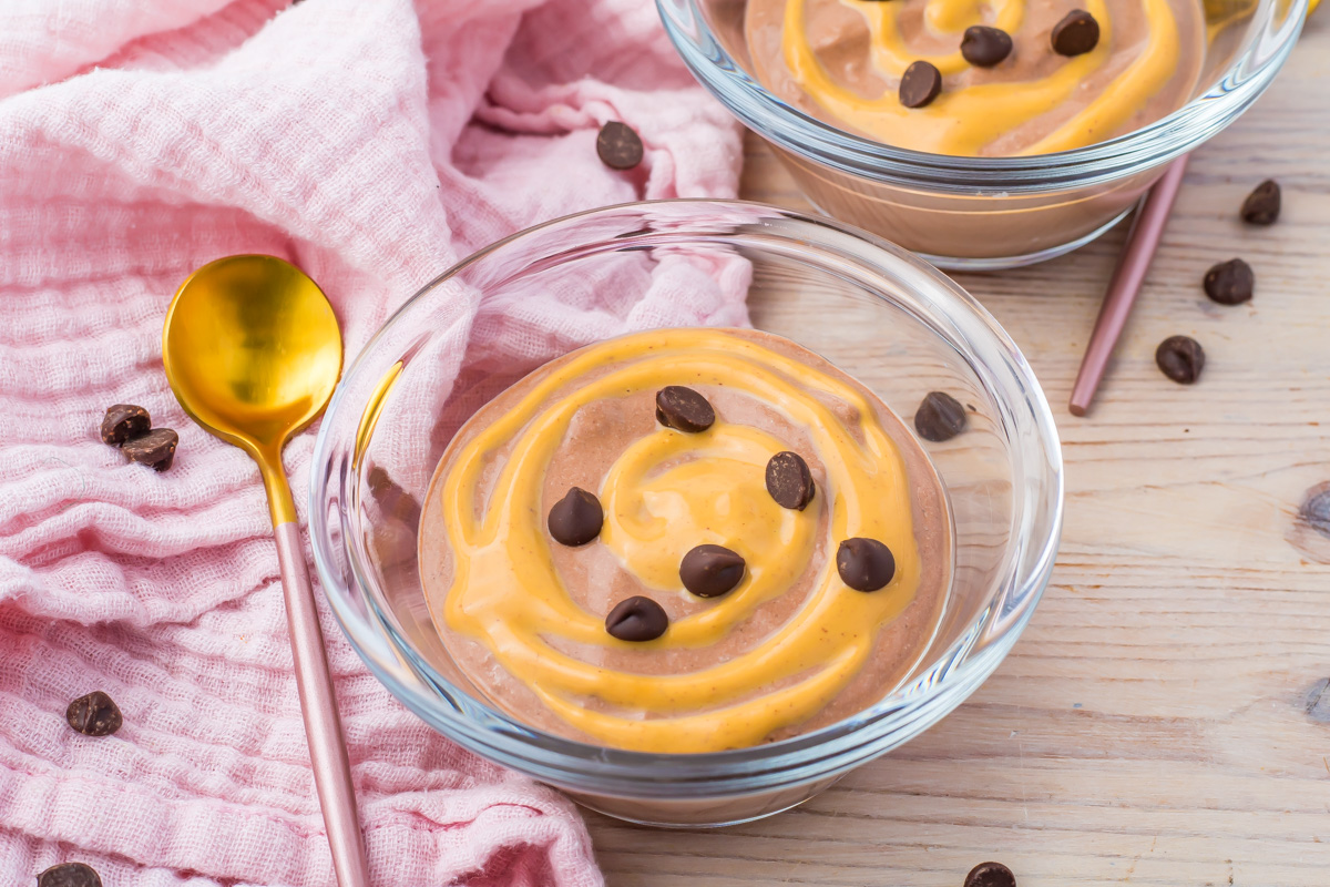 Glass bowls of swirled chocolate and peanut butter cottage cheese mousse topped with chocolate chips. Spoons with pink handles lie beside, on a wooden surface with a pink cloth.