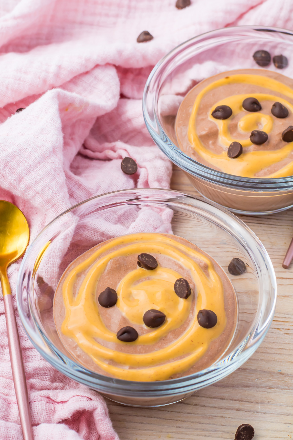 Two glass bowls of cottage cheese mousse with a peanut butter swirl and chocolate chips sit on a wooden surface, accompanied by a pink cloth and pink spoon.