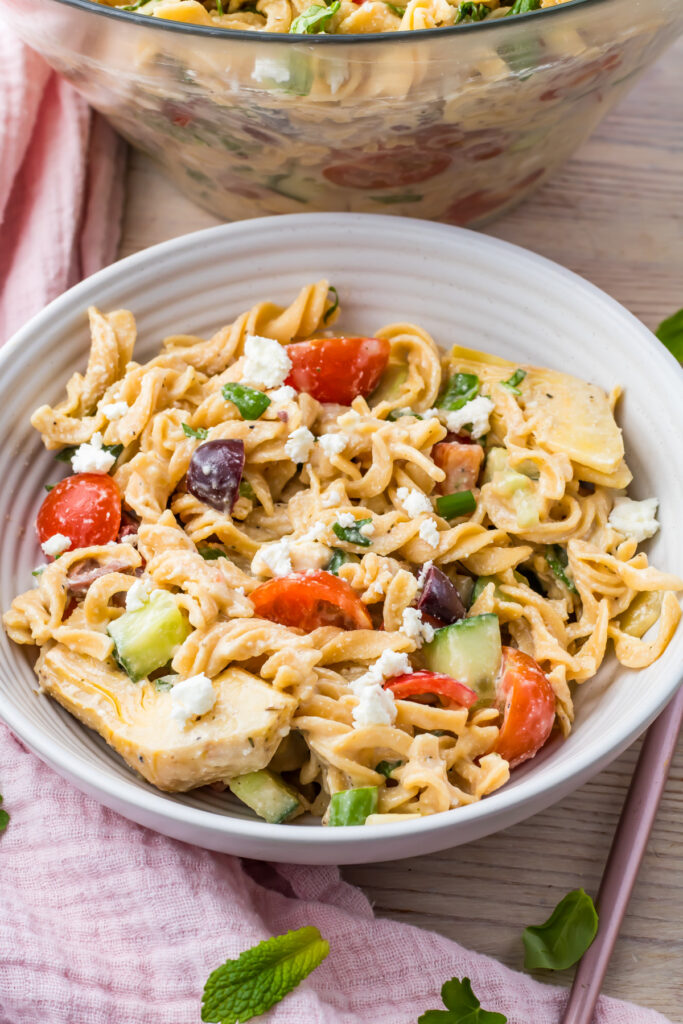 A bowl of high-protein pasta salad with cherry tomatoes, cucumbers, olives, feta cheese, and fresh herbs sits on a wooden table beside a pink cloth.