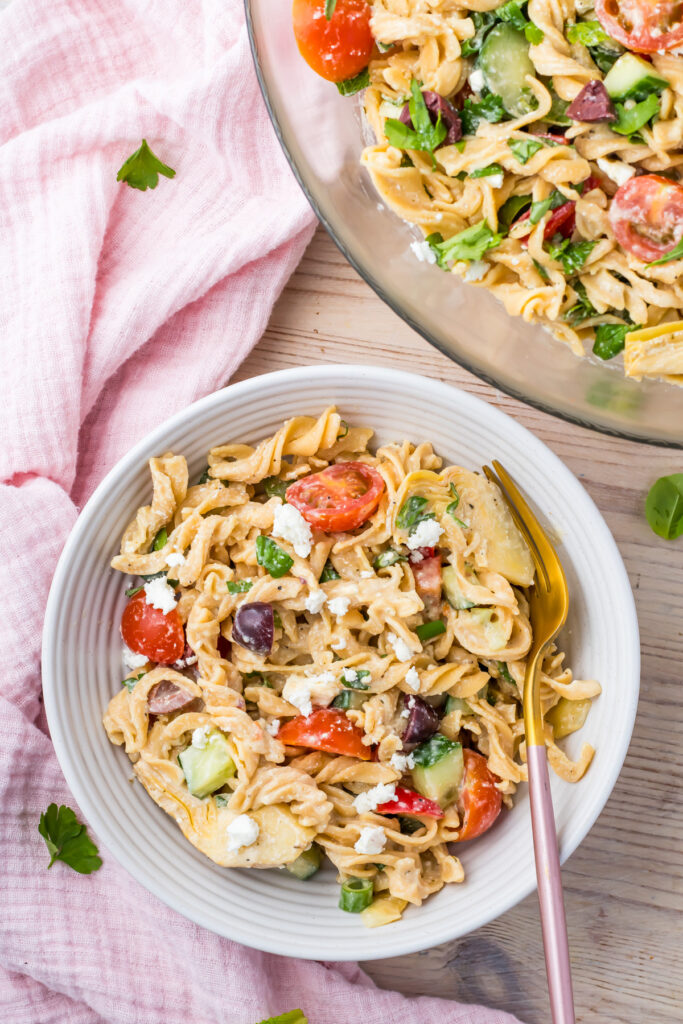A high-protein pasta salad featuring cherry tomatoes, cucumbers, olives, artichokes, parsley, and feta cheese sits in a bowl next to a pink cloth, with a fork ready on the side.