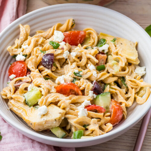A high-protein pasta salad featuring cherry tomatoes, cucumbers, olives, feta cheese, and artichoke hearts is elegantly served in a white dish on a wooden table.