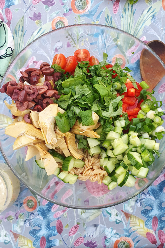 A glass bowl filled with chopped ingredients: cucumbers, red bell peppers, cherry tomatoes, olives, herbs, and artichokes sits on a floral-patterned tablecloth.