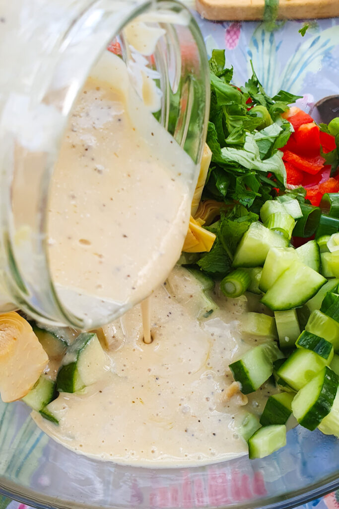 High-protein dressing is being poured from a glass jar onto a salad with cucumbers, herbs, artichokes, and red peppers.