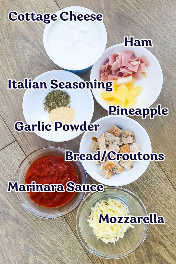 Overhead view of bowls containing cottage cheese, ham, Italian seasoning, pineapple, garlic powder, croutons, marinara sauce, and mozzarella on a wooden surface.