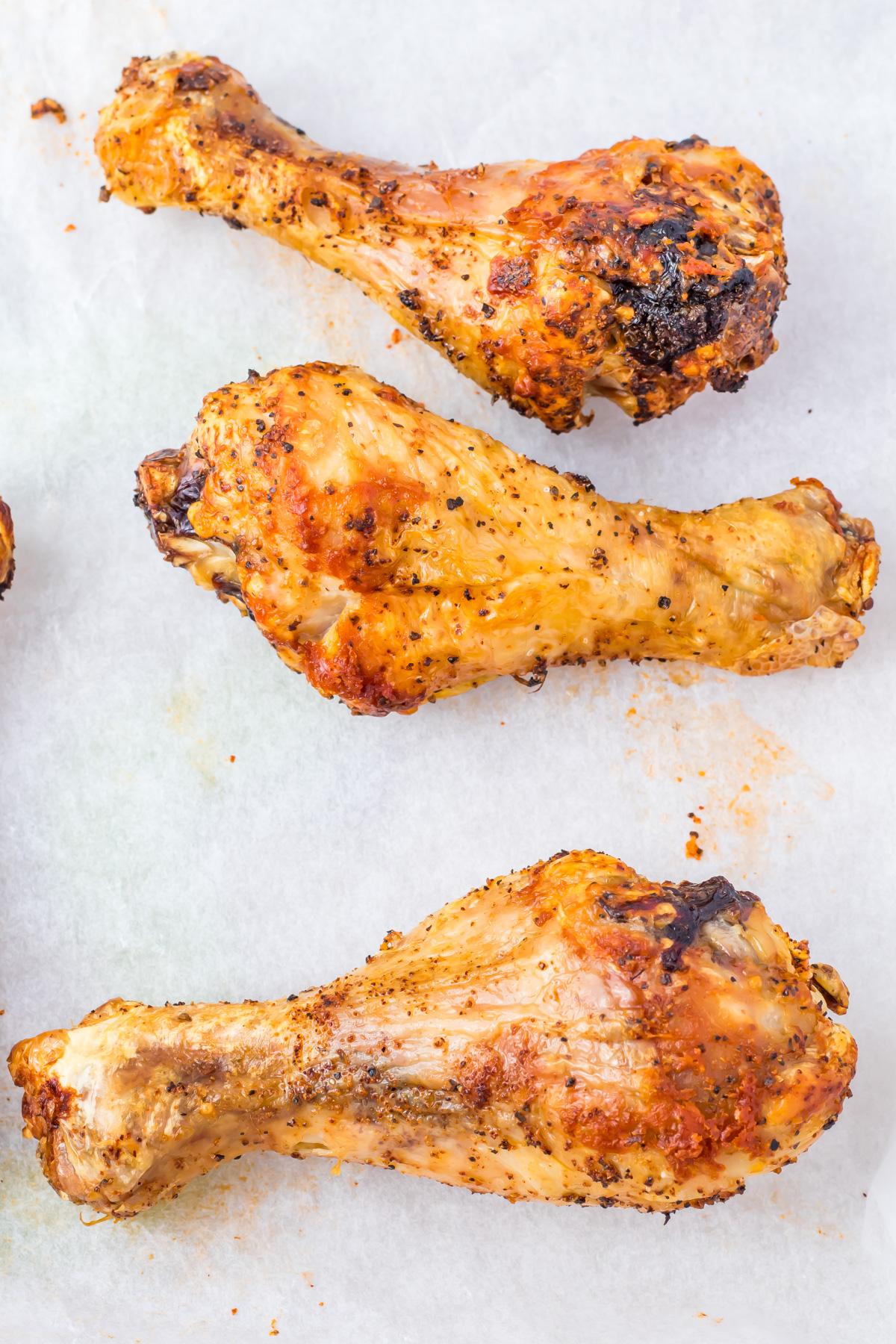Three oven-baked chicken drumsticks, seasoned with herbs and spices, rest invitingly on a white parchment paper surface.