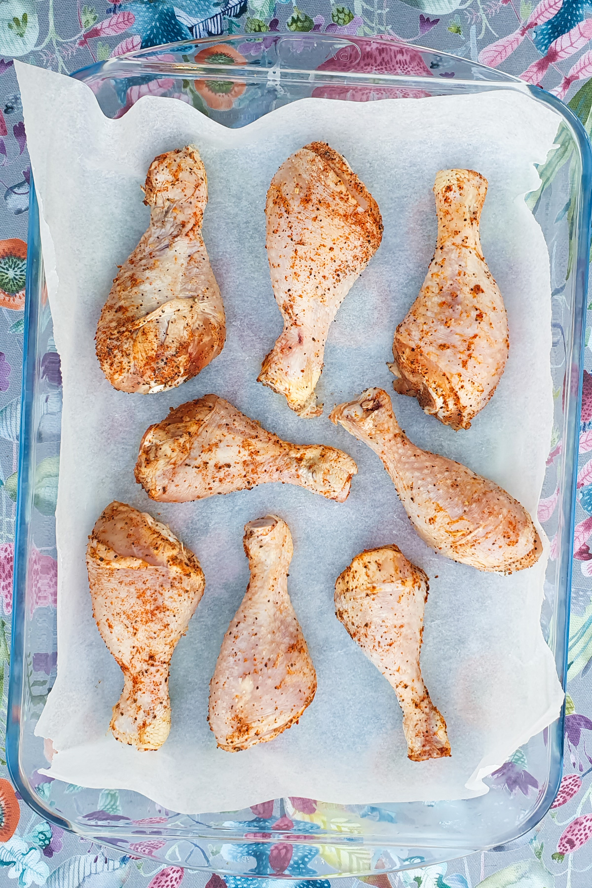 Eight seasoned oven baked drumsticks are expertly arranged on parchment paper in a glass baking dish, ready for cooking.