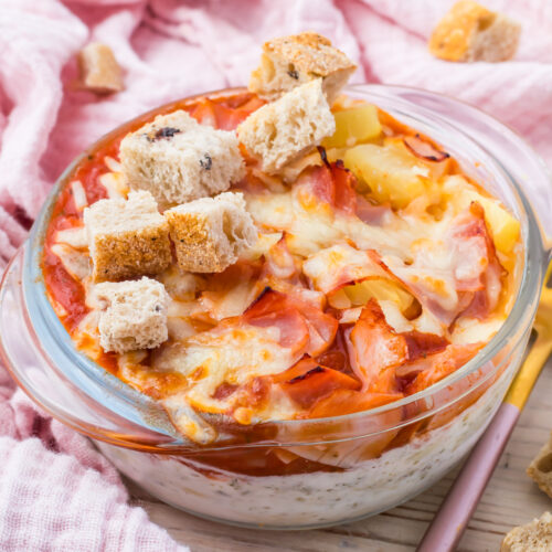 A cottage cheese pizza bowl topped with cheese, ham, pineapple, and croutons. The appetizing display rests on a pink cloth.