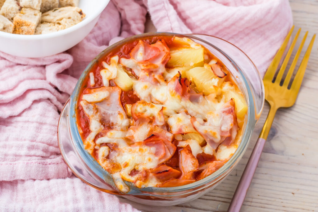 A cottage cheese pizza bowl, including melted cheese, ham, and pineapple, is placed on a pink cloth. A golden fork and a bowl of bread cubes are beside it.