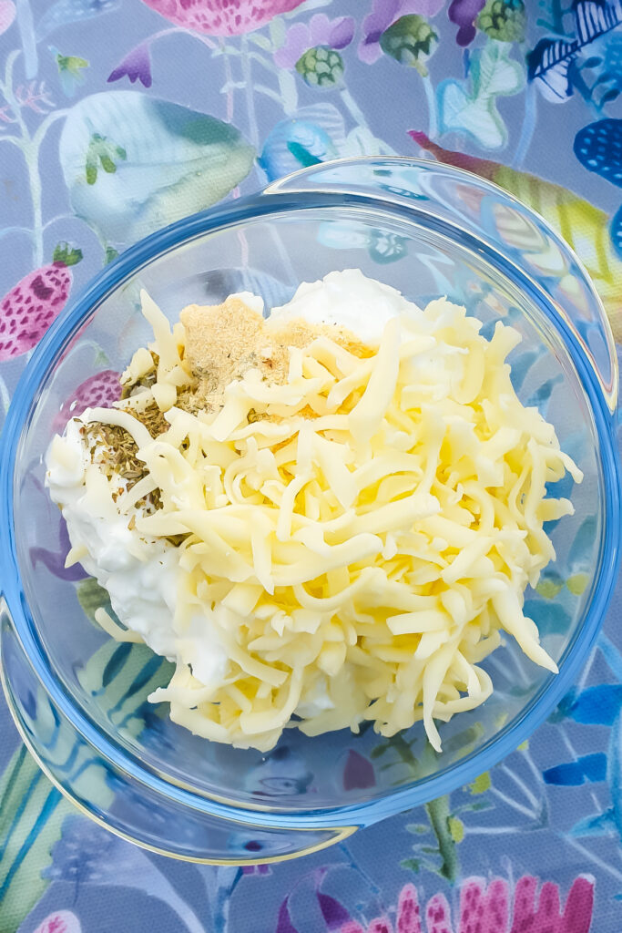 A glass bowl brimming with shredded cheese, cottage cheese, and seasonings rests on a floral-patterned tablecloth.