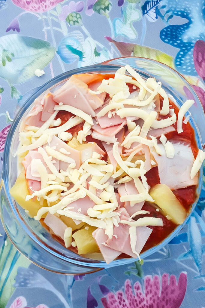 A bowl of pizza ingredients, featuring diced ham, shredded cheese, pineapple, and tomato sauce, rests on a floral tablecloth. A dollop of cottage cheese adds a creamy twist to this classic mix.